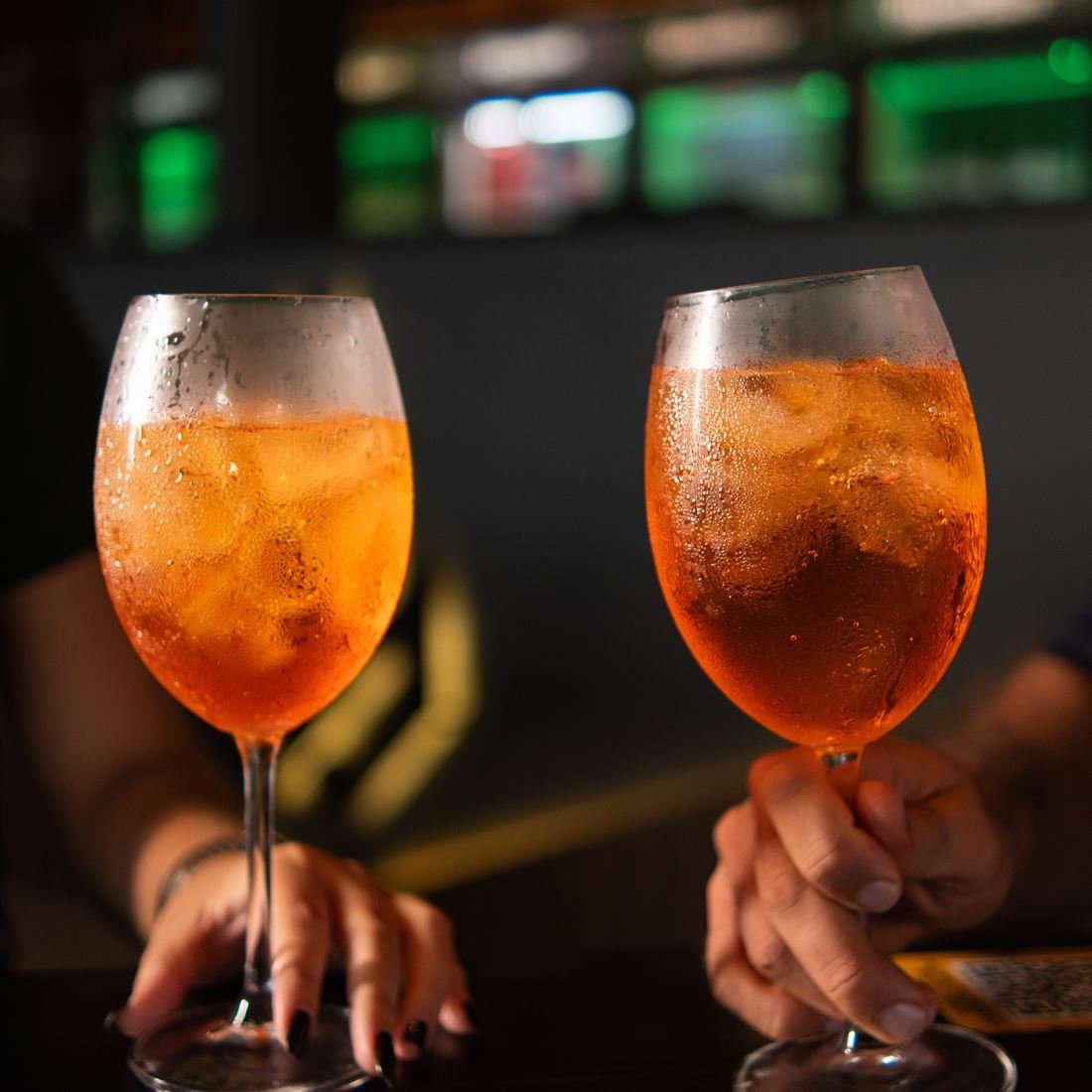 Duas taças de drink laranja com gelo, seguradas no balcão do bar à noite